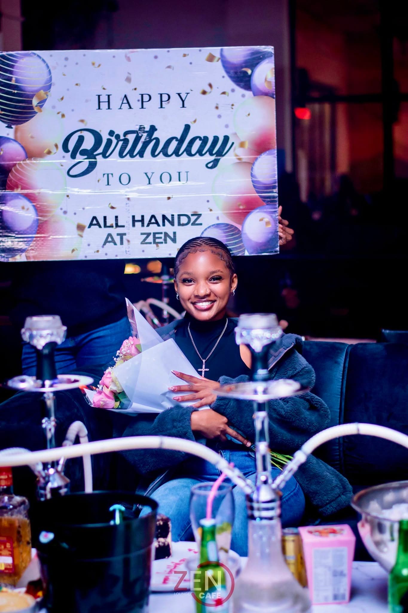A guest beams while holding a 'Happy Birthday' sign, hookah pipes in the foreground.