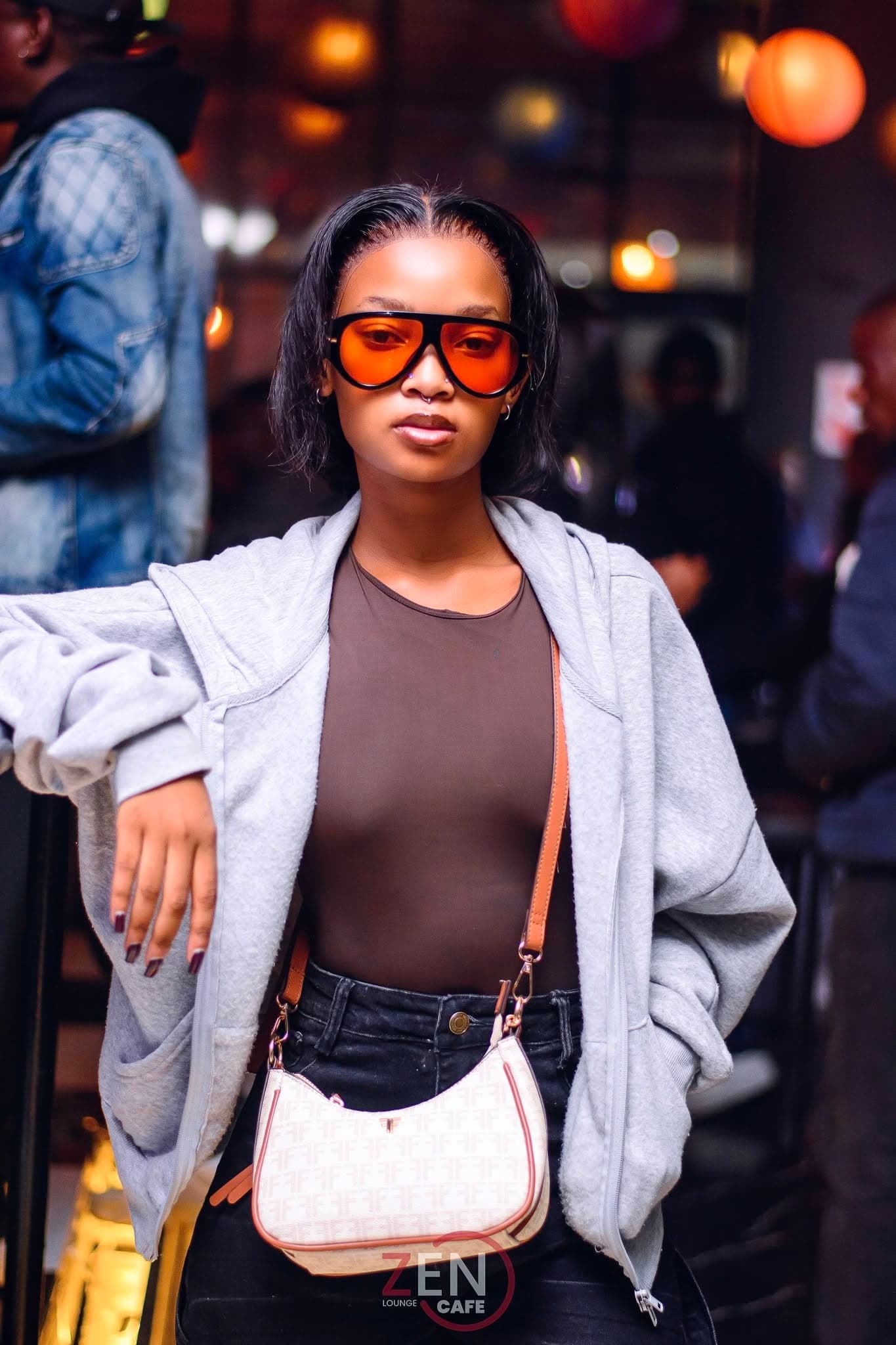 A guest in tinted sunglasses and an oversized hoodie poses at Zen Cafe Lounge, warm lantern light glowing behind her.